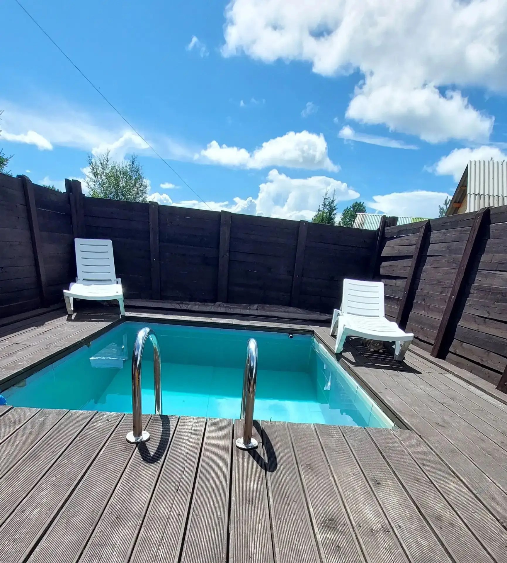 Private pool with a ladder and lounge chairs on a wooden deck, surrounded by a high fence.